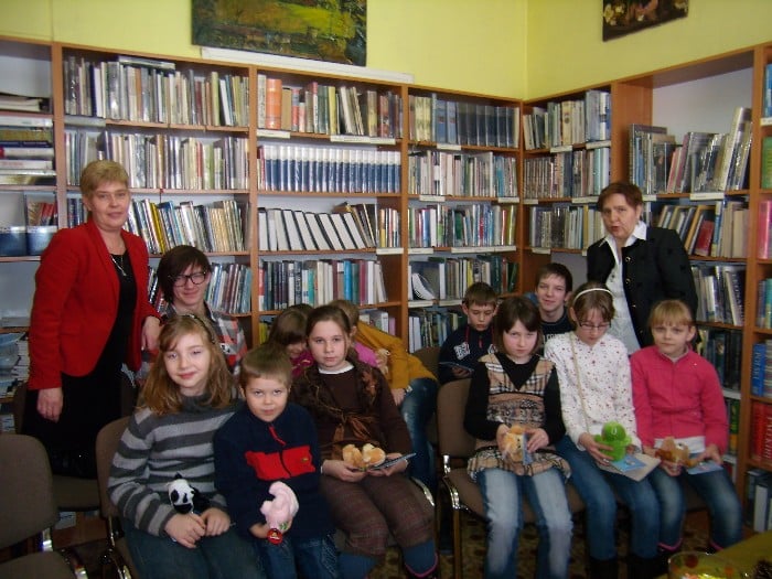 Przy regałach z książkami siedzą dzieci po bokach dwie bibliotekarki. 
