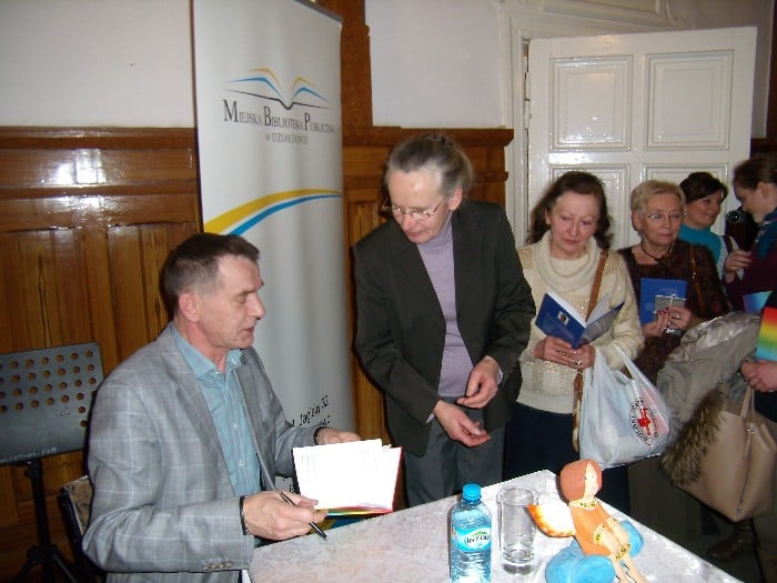 Przy stoliku siedzi autor Krzysztof Tabaczka i podpisuje czytelnikom książki.