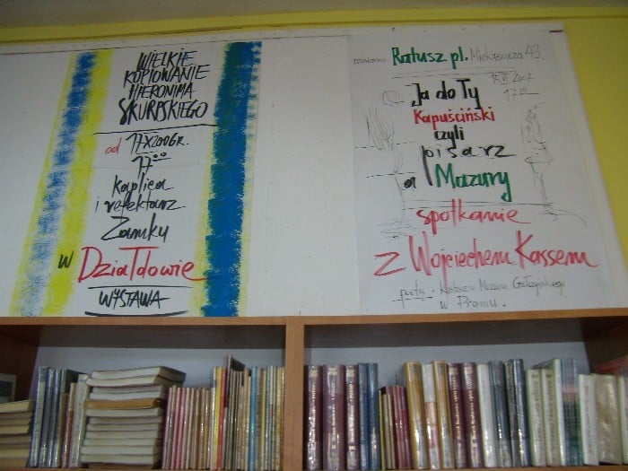 Wnętrze biblioteki, na ścianie wiszą plakaty Wiesława Wachowskiego.