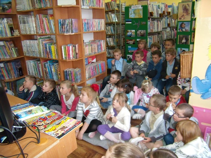 Wnętrze biblioteki, grupa dzieci siedzi między regałami na wykładzinie zebrała się z okazji Jubileuszu 10-lecia kampanii społecznej „Cała Polska czyta dzieciom”.