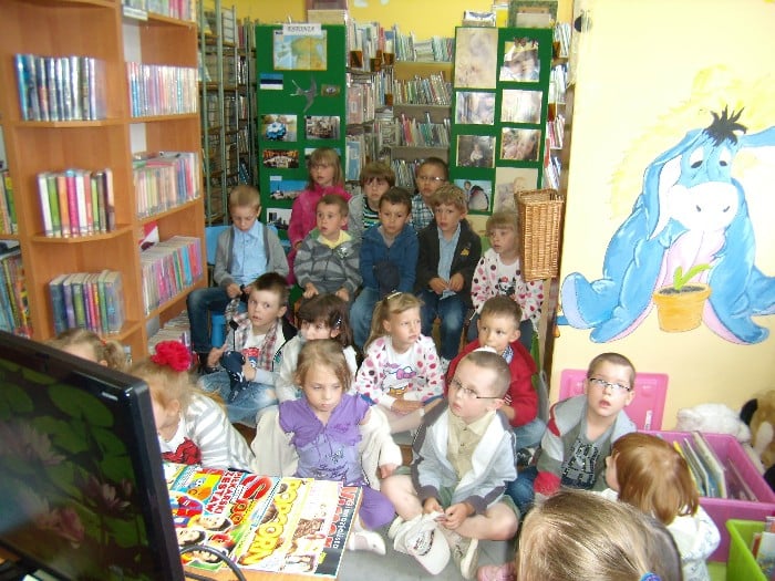 Wnętrze biblioteki, grupa dzieci siedzi między regałami na wykładzinie zebrała się z okazji Jubileuszu 10-lecia kampanii społecznej „Cała Polska czyta dzieciom”.