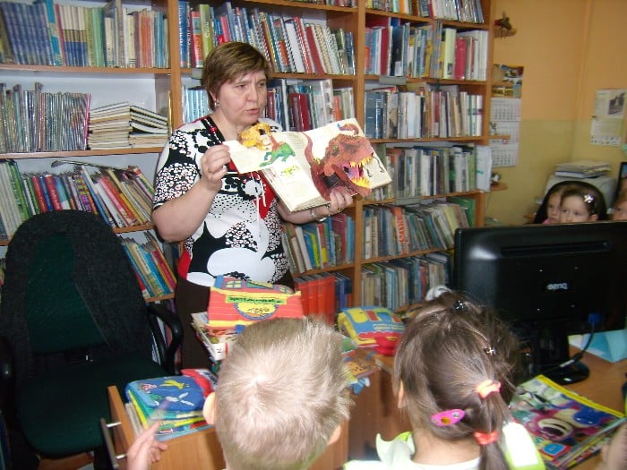 Wnętrze biblioteki, grupa  5-latków z Przedszkola nr 4 w Działdowie na lekcji bibliotecznej, pani bibliotekarka pokazuje dzieciom książki.