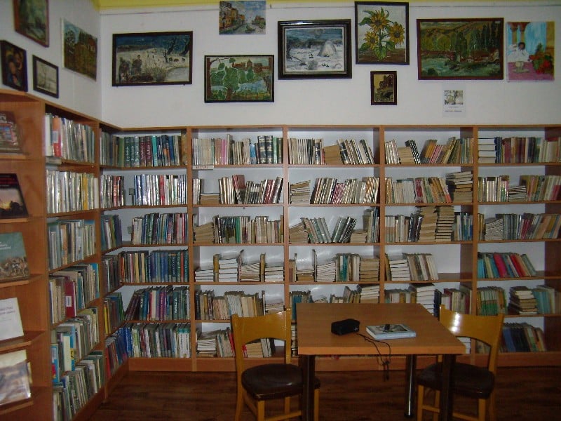 Wnętrze biblioteki, na środku stoi stół z krzesłami oraz regały z książkami.
