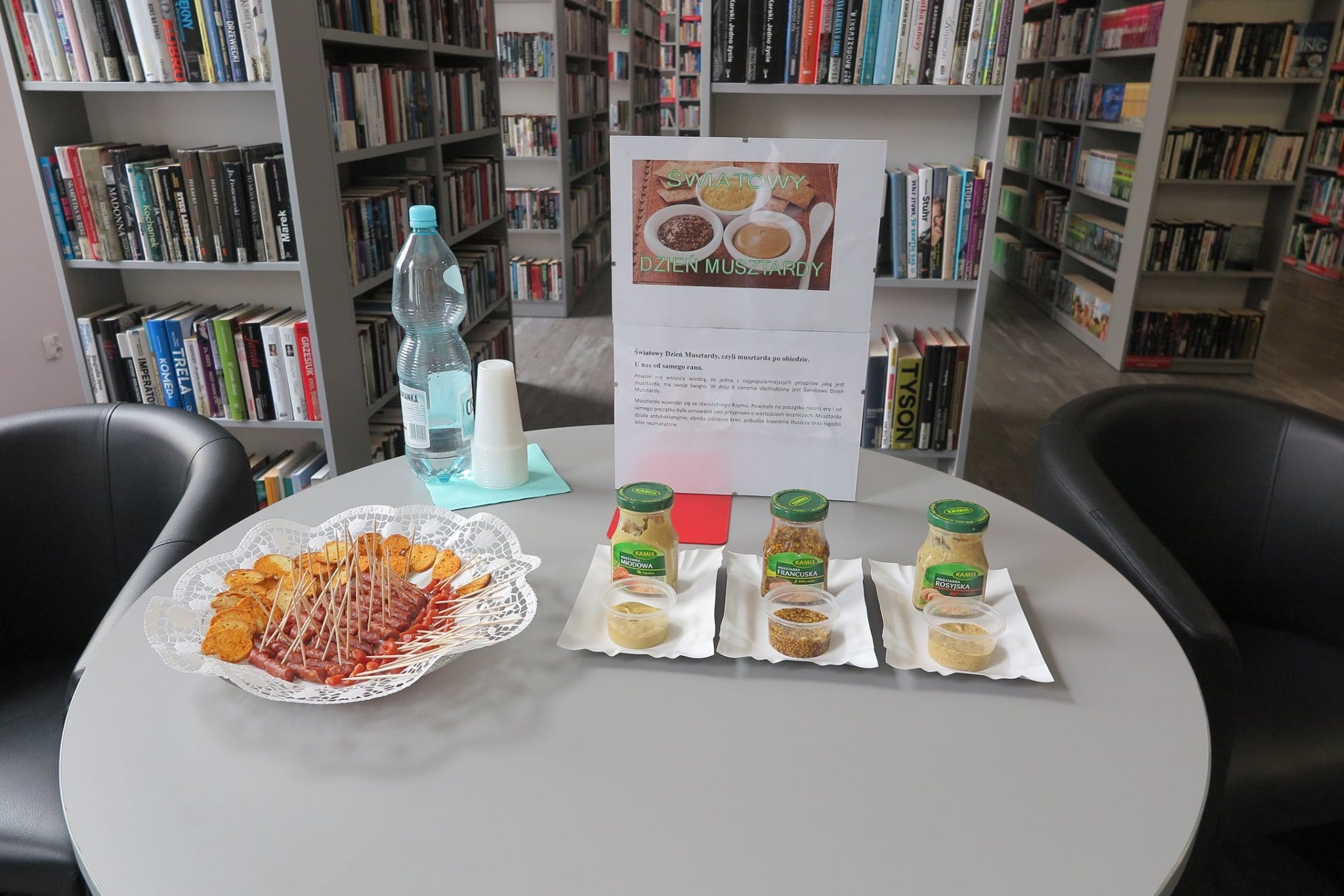 Wnętrze biblioteki. Na stoliku plakat ''Światowy Dzień Musztardy'' do degustacji stoją trzy słoiczki musztardy, na talerzu mini kiełbaski oraz woda.