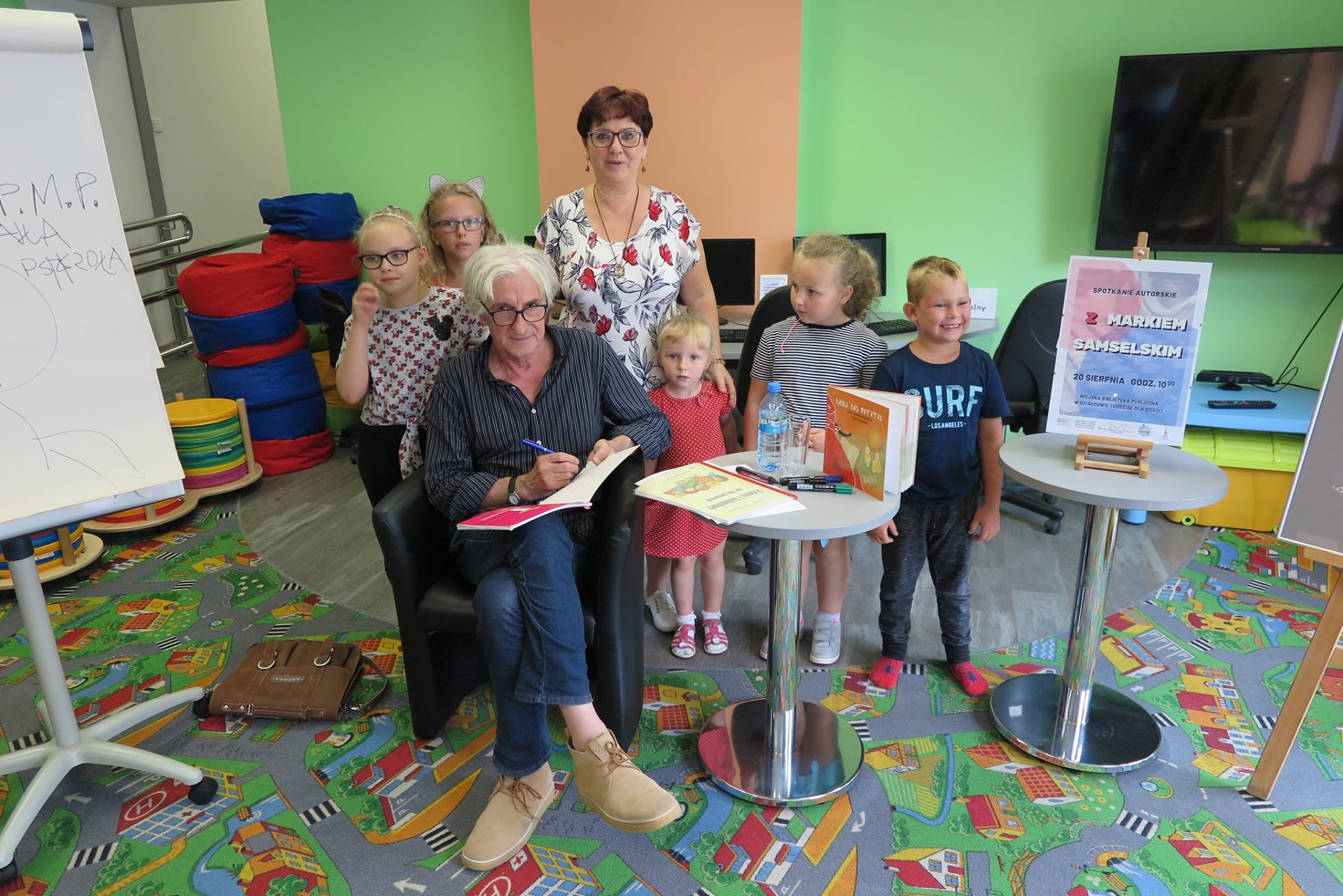 Wnętrze biblioteki, oddział dla dzieci. Na fotelu autor Marek Samselski obok pięcioro dzieci i opiekun.