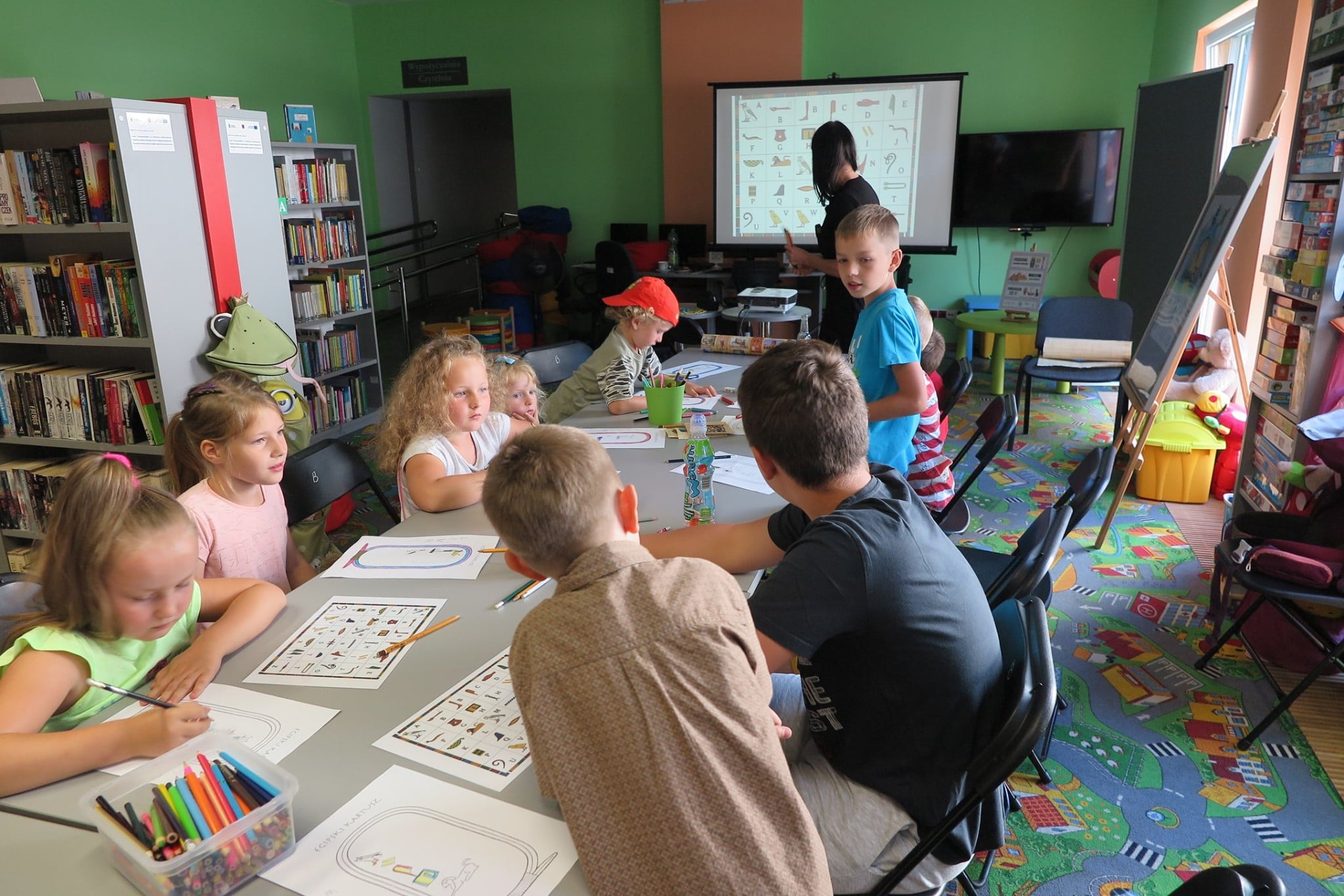 Wnętrze biblioteki, oddział dla dzieci. Warsztaty archeologiczne prowadzi z dziećmi Anna Jaklewicz. Na stole kredki,długopisy i kartki. Dzieci wykonują prace plastyczne. 