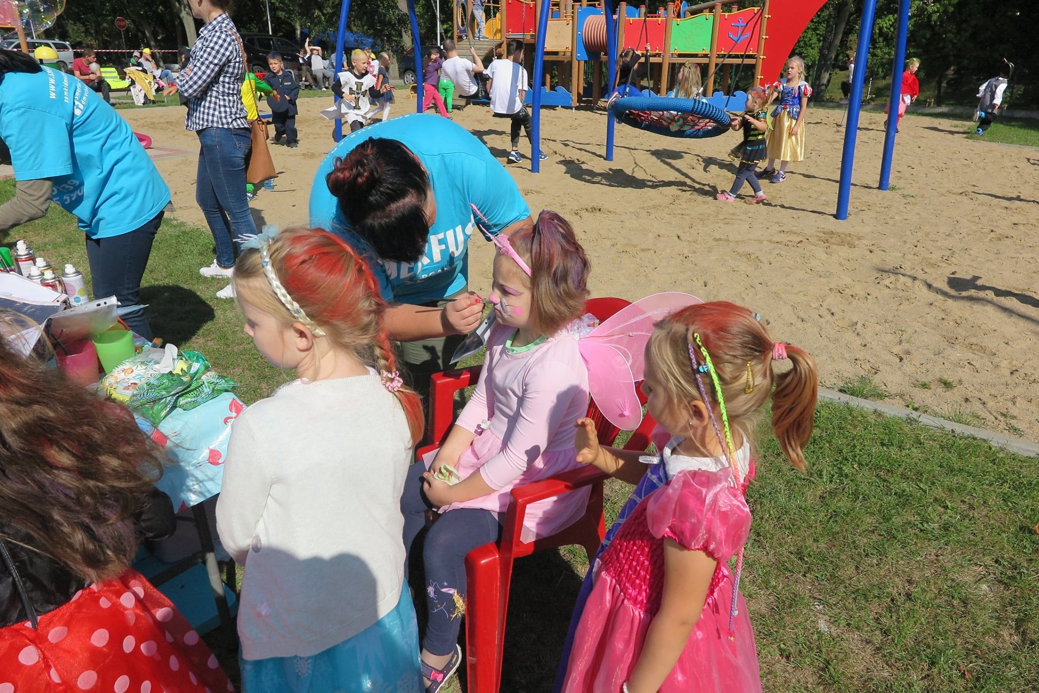 Grupa dzieci na placu zabaw obok biblioteki maluje twarze z okazji ''Parady Bajkowych Postaci''.