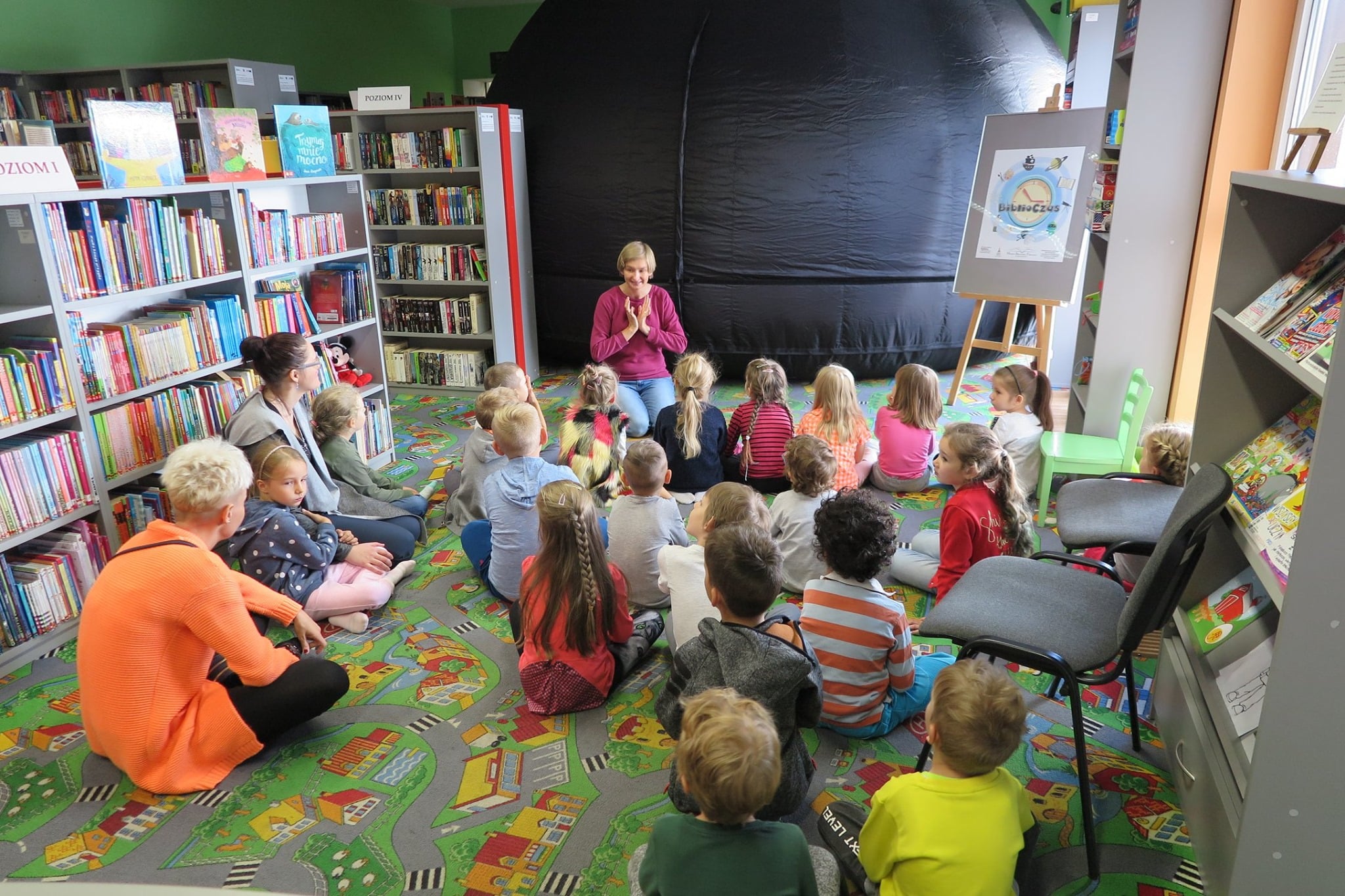 Wnętrze biblioteki, duża grupa dzieci siedzi na kolorowej wykładzinie, w tle mobilne planetarium. 