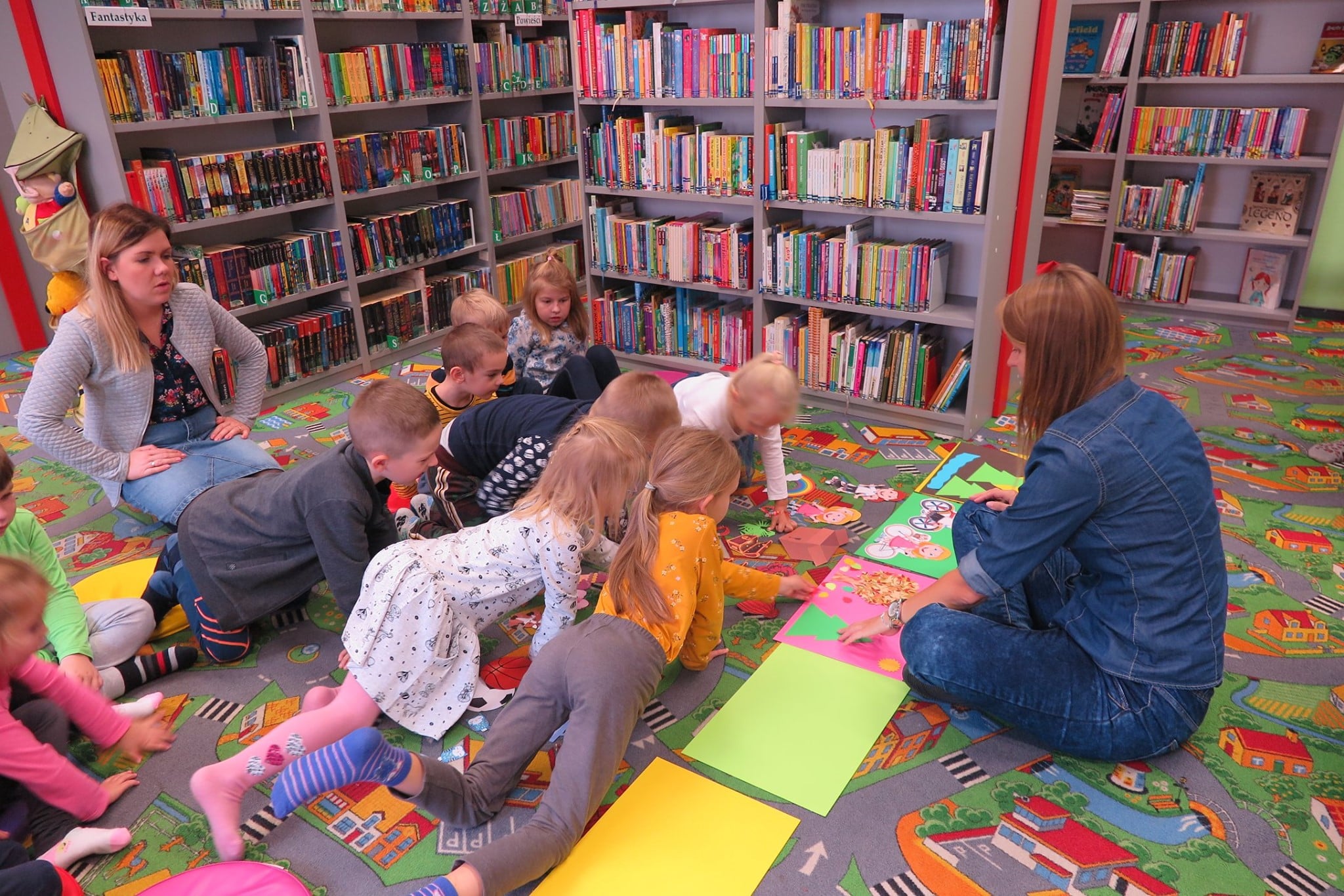 Wnętrze biblioteki ,oddział dla dzieci. Dzieci siedzą na pufach i oglądają swoje prace plastyczne. 