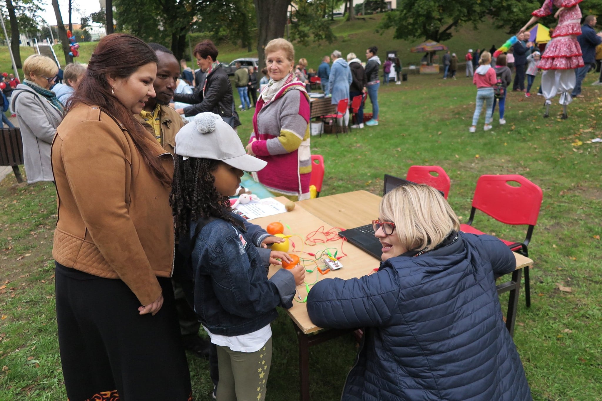 W parku przy stoliku bibliotekarka Dorota Kusik oraz rodzic z dzieckiem grają w interaktywną grę z użyciem płytek makey-makey z okazji pomarańczowej niedzieli cittaslow.