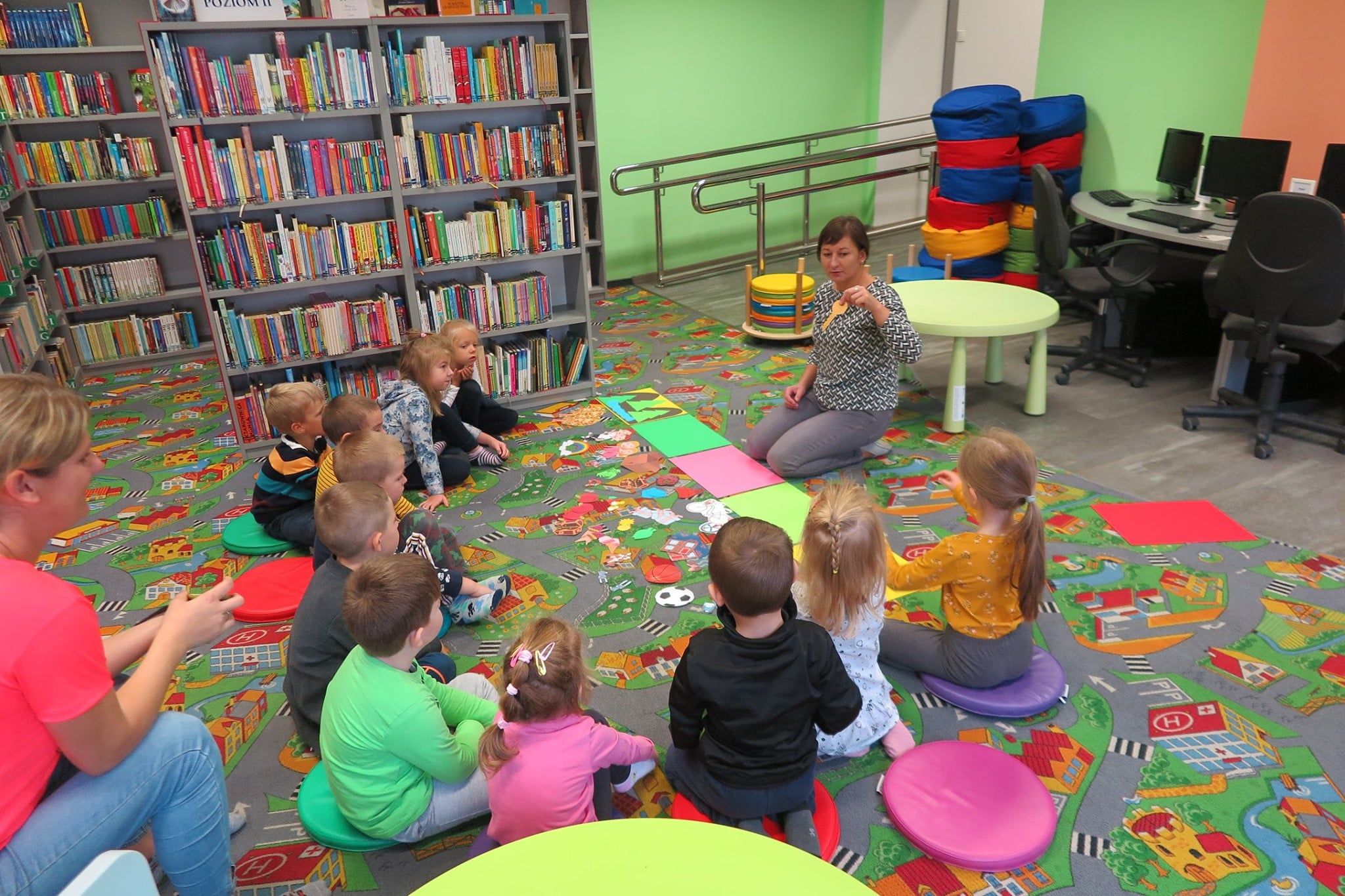 Wnętrze biblioteki ,oddział dla dzieci. Dzieci siedzą na pufach i biorą udział w  warsztatach literacko-plastycznych. Zajęcia prowadzi bibliotekarka Ania Wiśniewska.