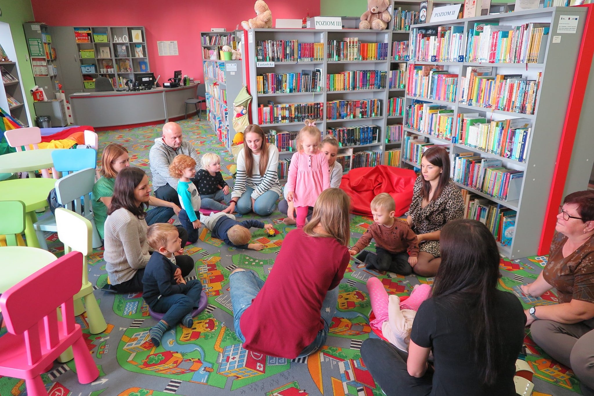 Wnętrze biblioteki (Klub Malucha) w kole siedzą dzieci wraz z opiekunami.