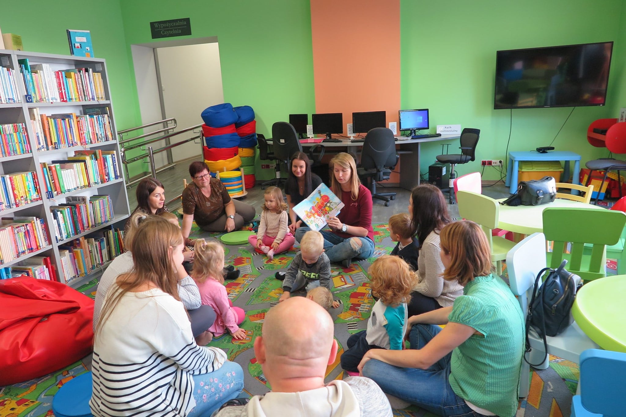 Wnętrze biblioteki (Klub Malucha) w kole siedzą dzieci wraz z opiekunami. Książkę ''Lokomotywa'' Juliana Tuwima czyta bibliotekarka Aleksandra Cybulska. 