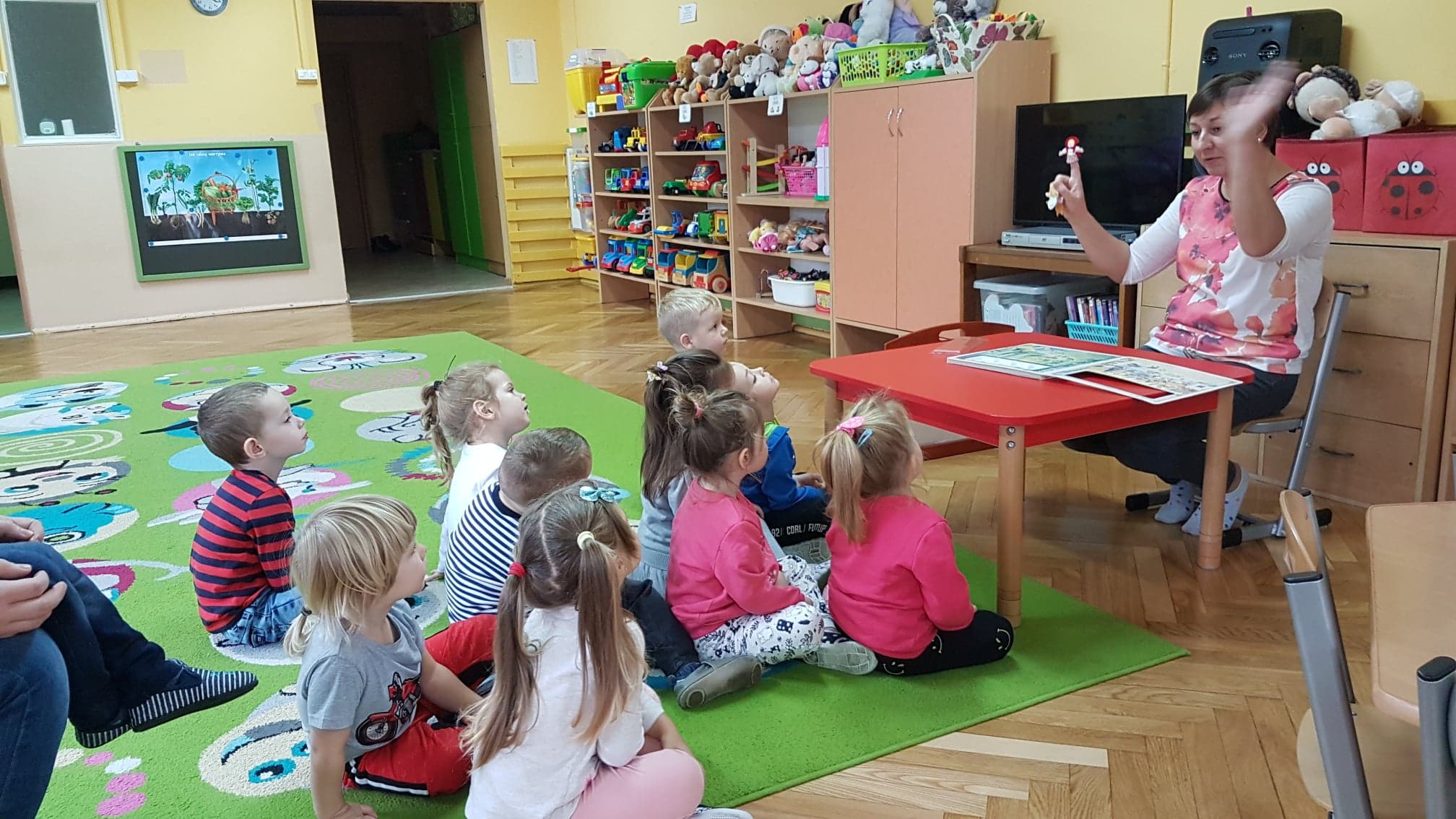 Wnętrze Przedszkole nr 4 na zielonej wykładzinie siedzą dzieci przy stoliku bibliotekarka Ania Wiśniewska czyta teatrzyk paluszkowy ,,Czerwony Kapturek''. 