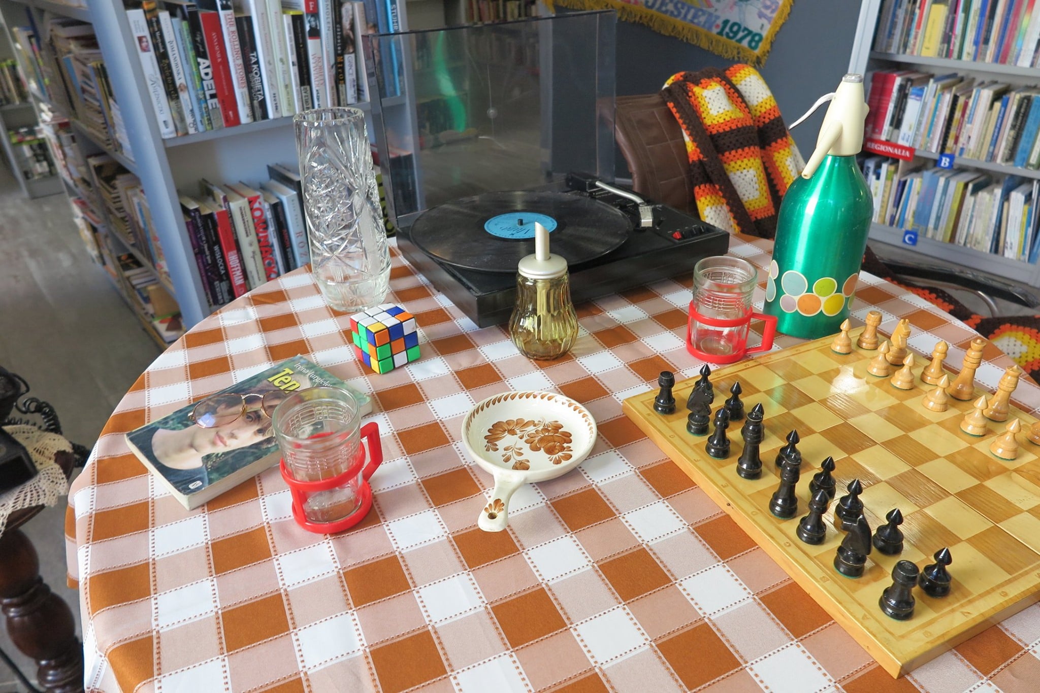 Na stoliku gramofon, szachy, charakterystyczne szklanki, wazon oraz naczynie z lat 80.