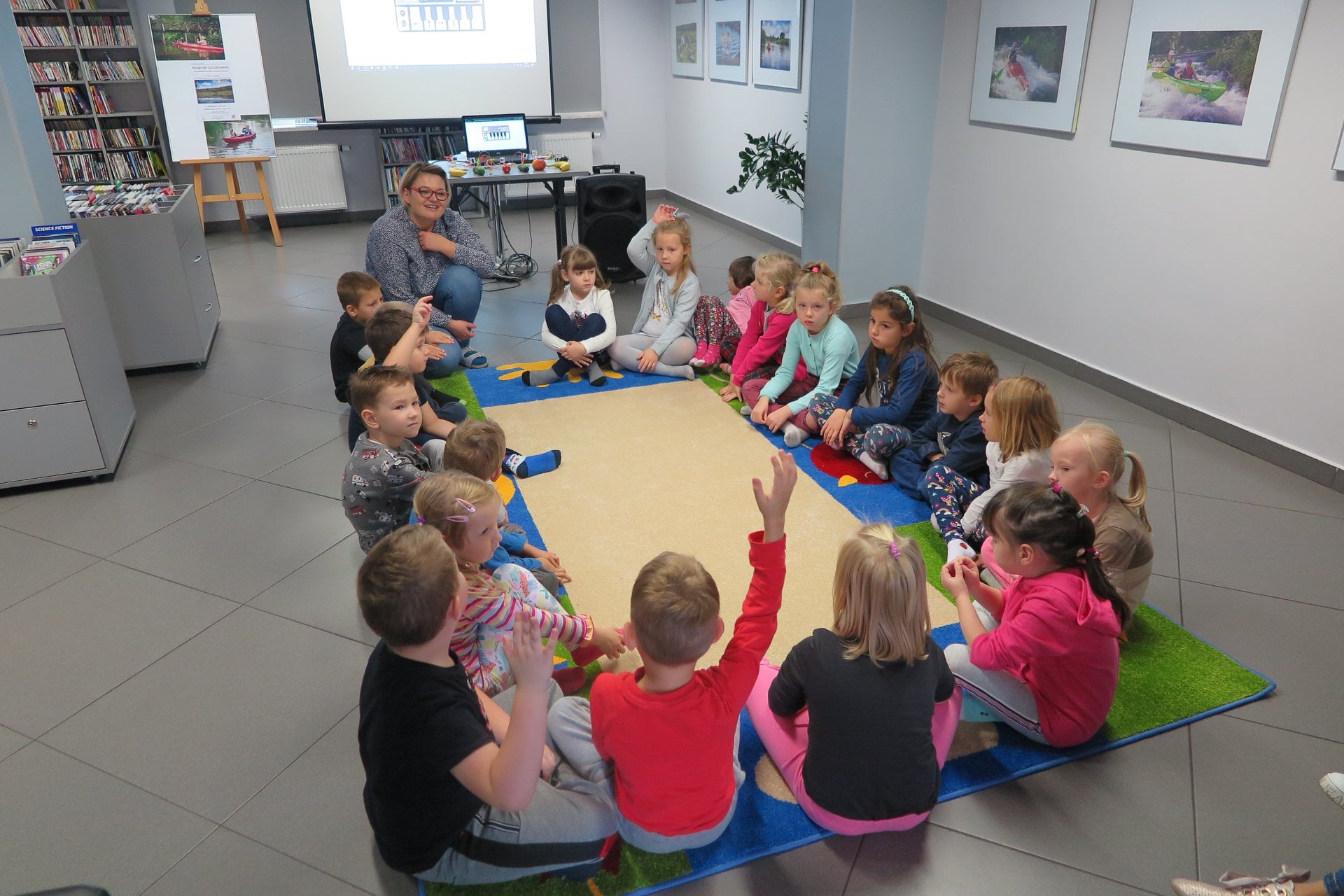 Wnętrze biblioteki,mediateka.Dzieci z przedszkola nr 3 grupa ''Motylki''  siedzą na kolorowym dywanie i biorą udział w warsztatach muzycznych. Zajęcia prowadzi bibliotekarka Dorota Kusik.