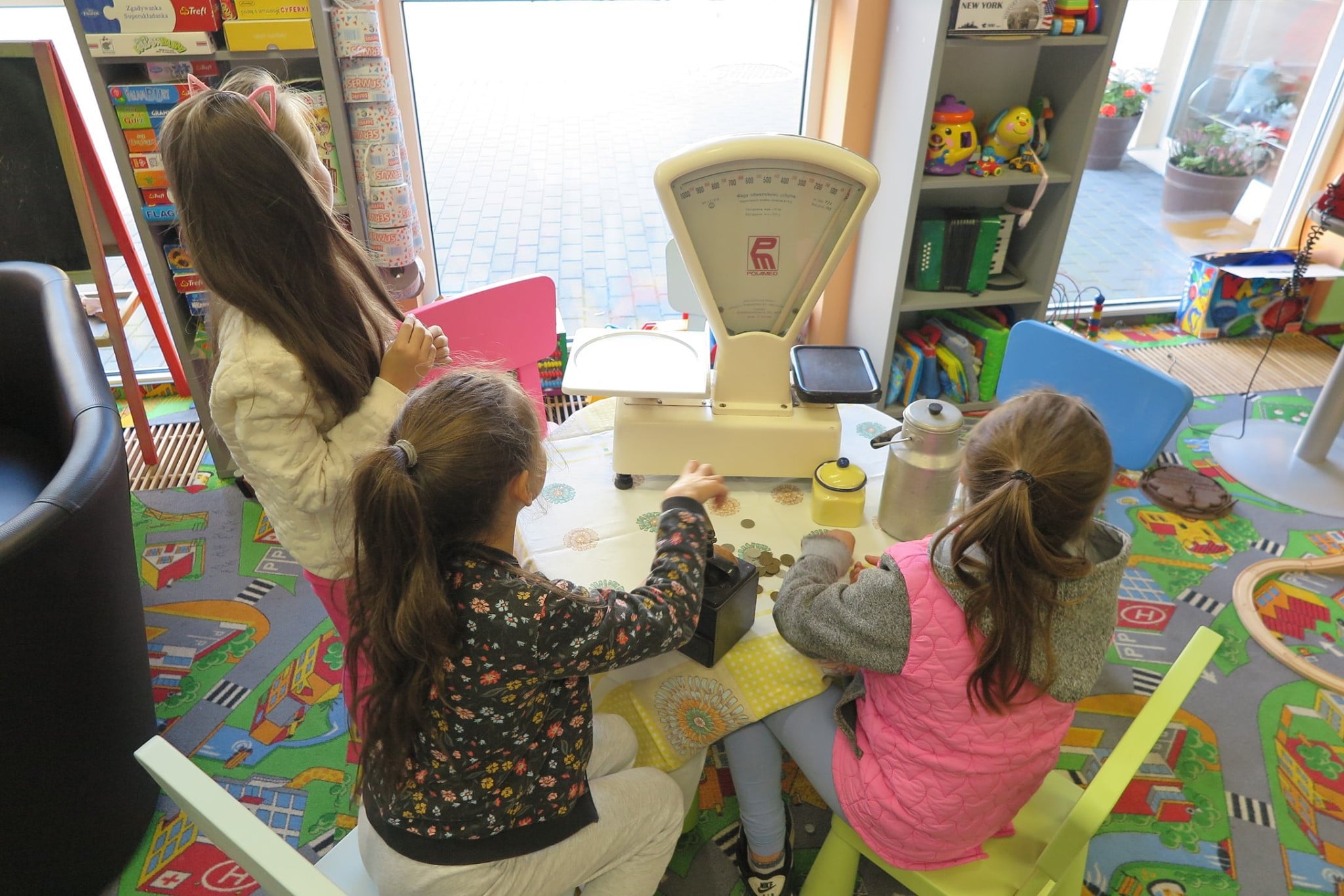 Wnętrze biblioteki,oddział dla dzieci. Przy stoliku siedzą trzy dziewczynki oglądają przedmioty z lat 80. Największy z nich waga.