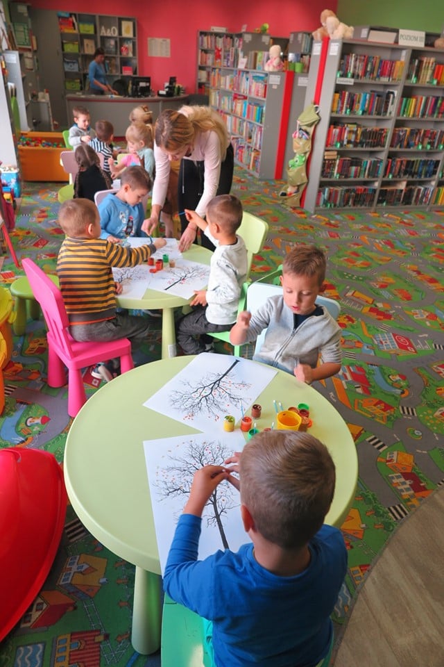 Wnętrze biblioteki, dzieci z Przedszkola nr 4  siedzą przy stolikach i malują farbami kolorowe jesienne drzewa.