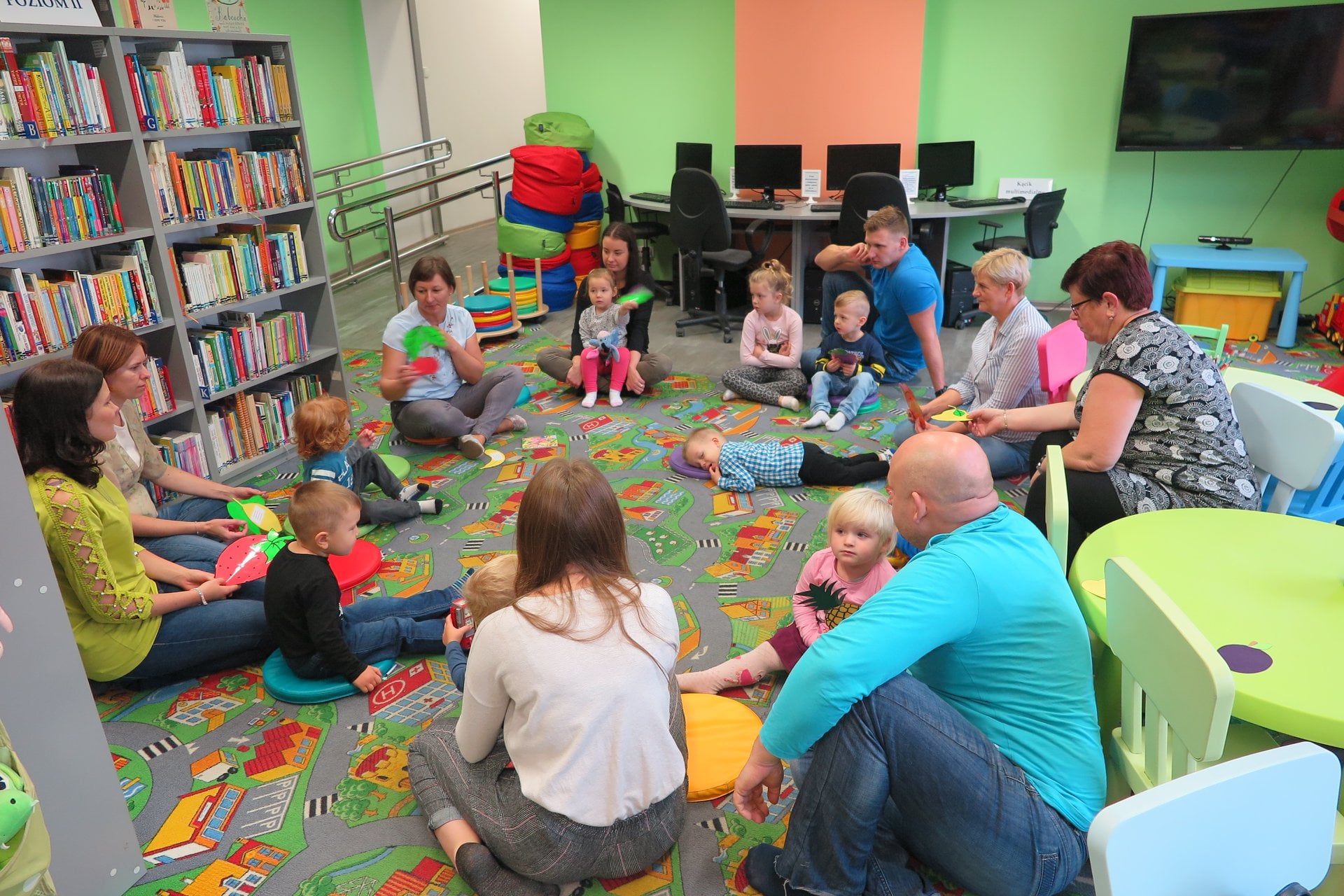 Wnętrze biblioteki, (Klub Malucha) Dzieci siedzą w kole wraz z opiekunami i poznają na zajęciach owoce. 
