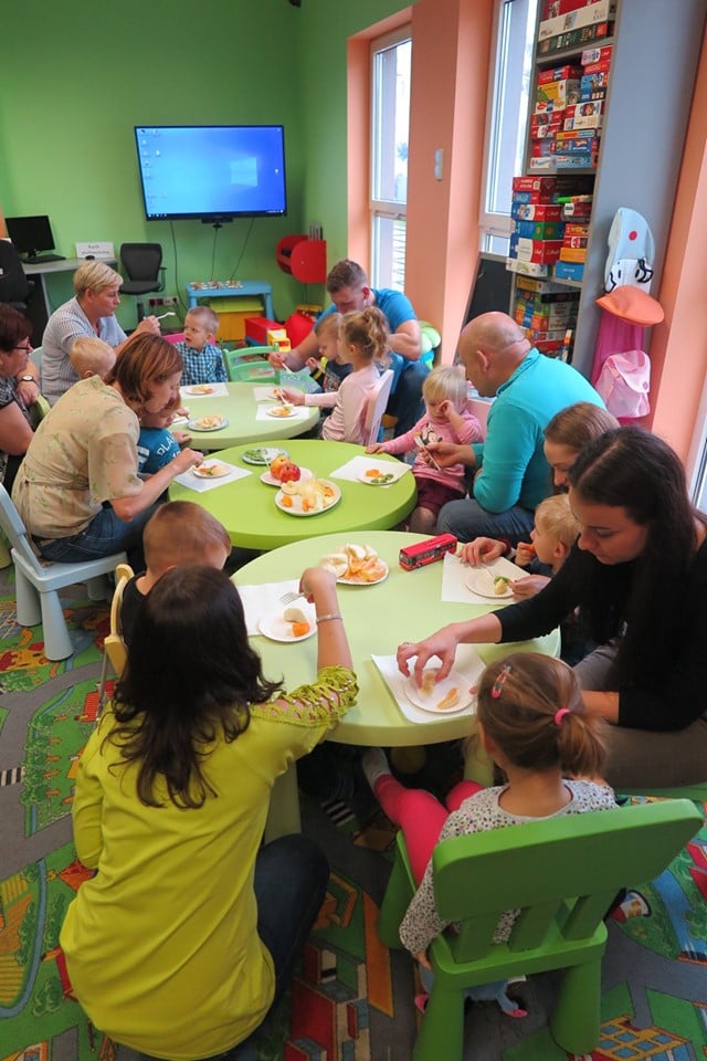 Wnętrze biblioteki, (Klub Malucha) dzieci wraz z opiekunami siedzą przy stolikach i degustują poznane owoce.