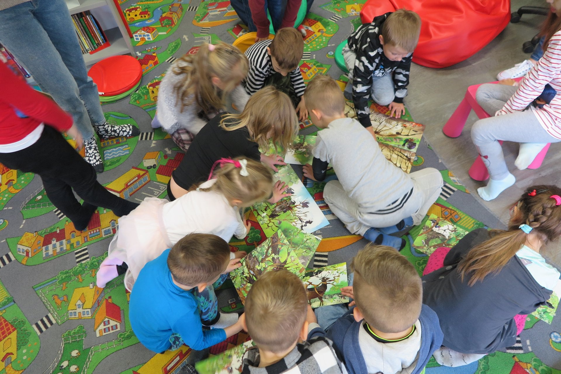 Wnętrze biblioteki. Na pufach siedzą dzieci ''Mądre sówki'' z przedszkola ''U Misia'' i układają książkę-puzzle z obrazkiem drzewa. 