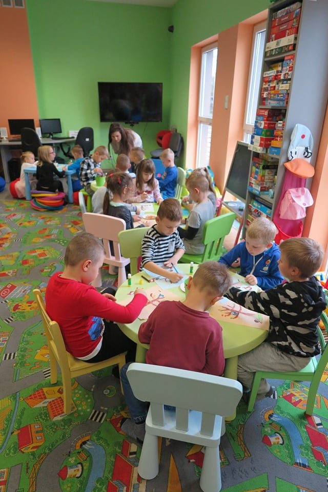 Wnętrze biblioteki. Dzieci z Przedszkola ''U Misia'' siedzą przy małych stolikach i wykonują prace jesienne drzewa z wcześniej przygotowanych elementów.