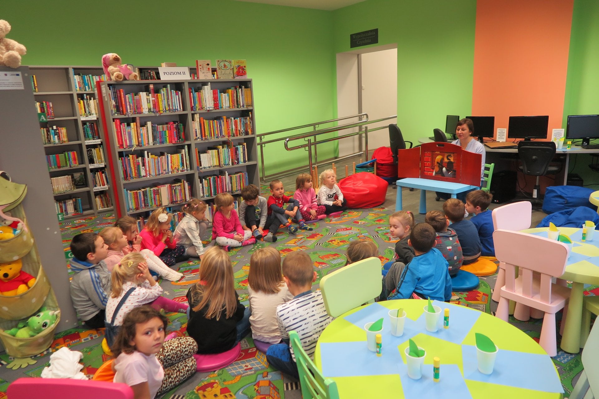 Wnętrze biblioteki. Na pufach siedzą dzieci z grupy III z Przedszkola nr 3. Dzieci słuchają teatrzyku Kamishibai ''O smoku wawelskim''który przedstawia bibliotekarka Ania Wiśniewska.