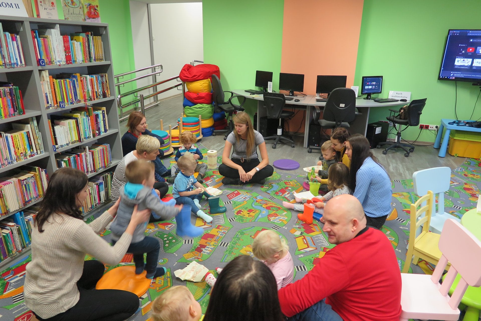 Wnętrze biblioteki (Klub Malucha), dzieci wraz z opiekunami prezentują swoje kolorowe kalosze. Zajęcia prowadzi bibliotekarka Aleksandra Cybulska.