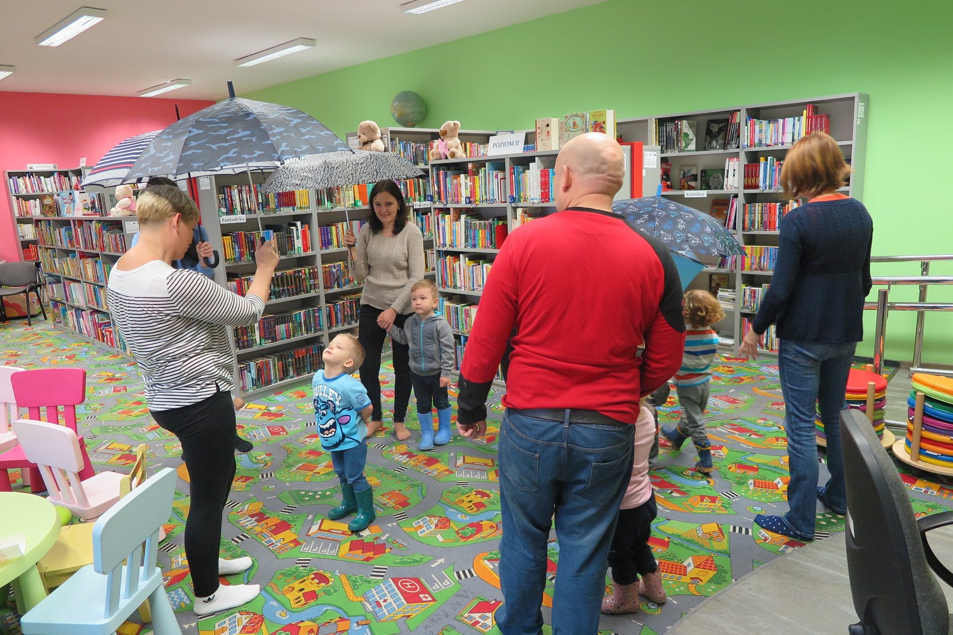 Wnętrze biblioteki (Klub Malucha), dzieci wraz z opiekunami tańczą do deszczowej piosenki trzymając w ręku parasolki. 