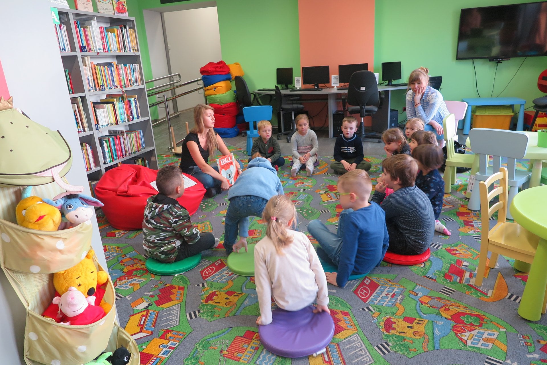 Wnętrze biblioteki. Dzieci z przedszkola nr.4 siedzą na pufach i słuchają książki pt. ''Koala, który się trzymał''. Zajęcia prowadzi bibliotekarka Aleksandra Cybulska. 