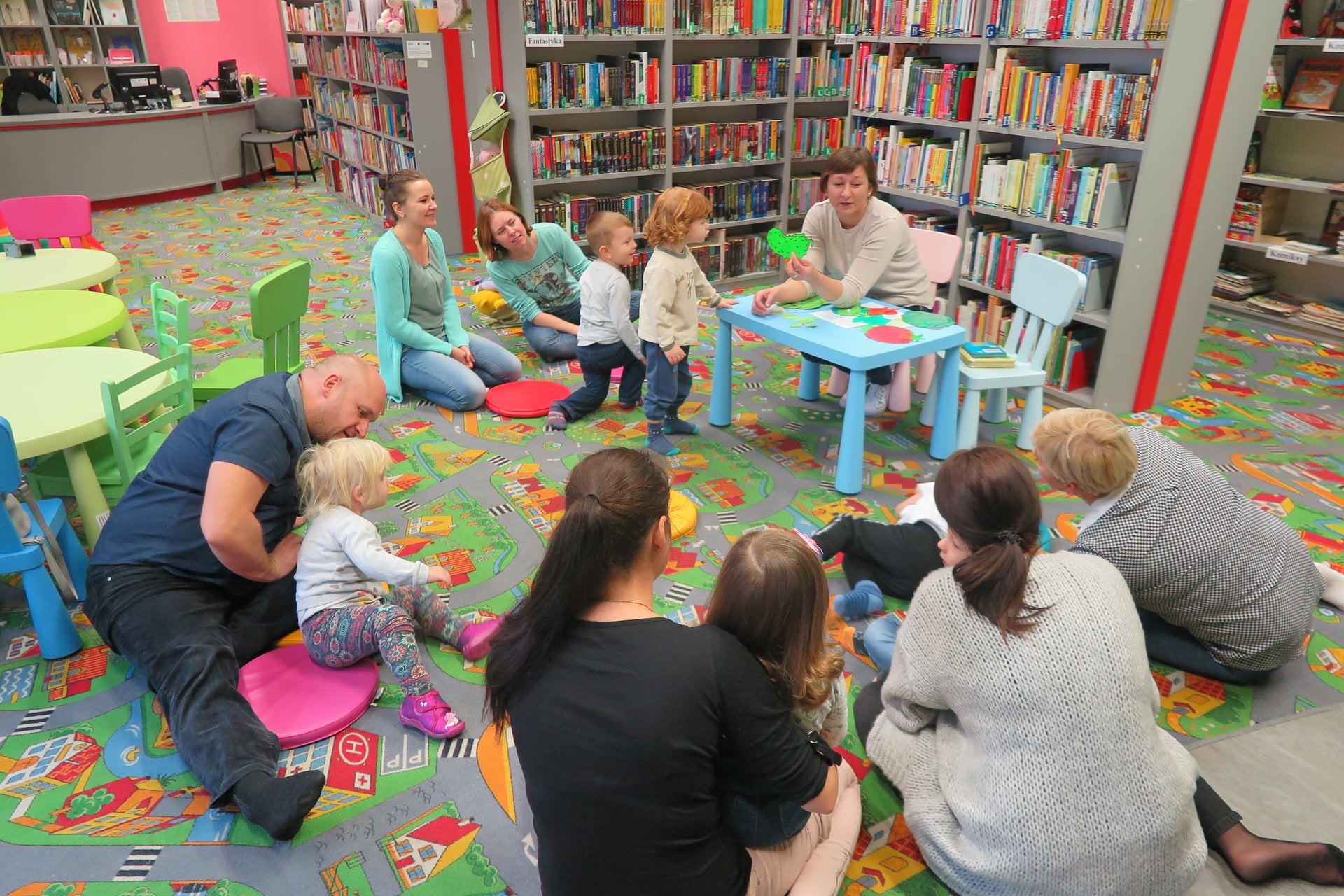 Wnętrze biblioteki (Klub Malucha) w małym kole na pufach siedzą dzieci wraz  z opiekunami. Zajęcia prowadzi bibliotekarka Ania Wiśniewska.