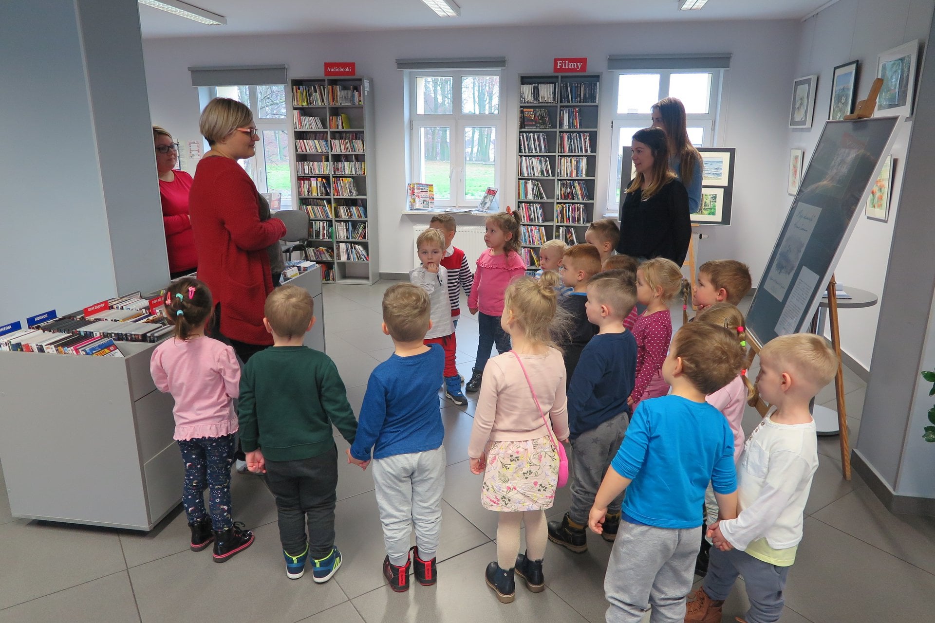 Wnętrze biblioteki. Dzieci z przedszkola ''U Misia'' zwiedzają mediatekę. O bibliotece opowiada bibliotekarka Dorota Kusik.  