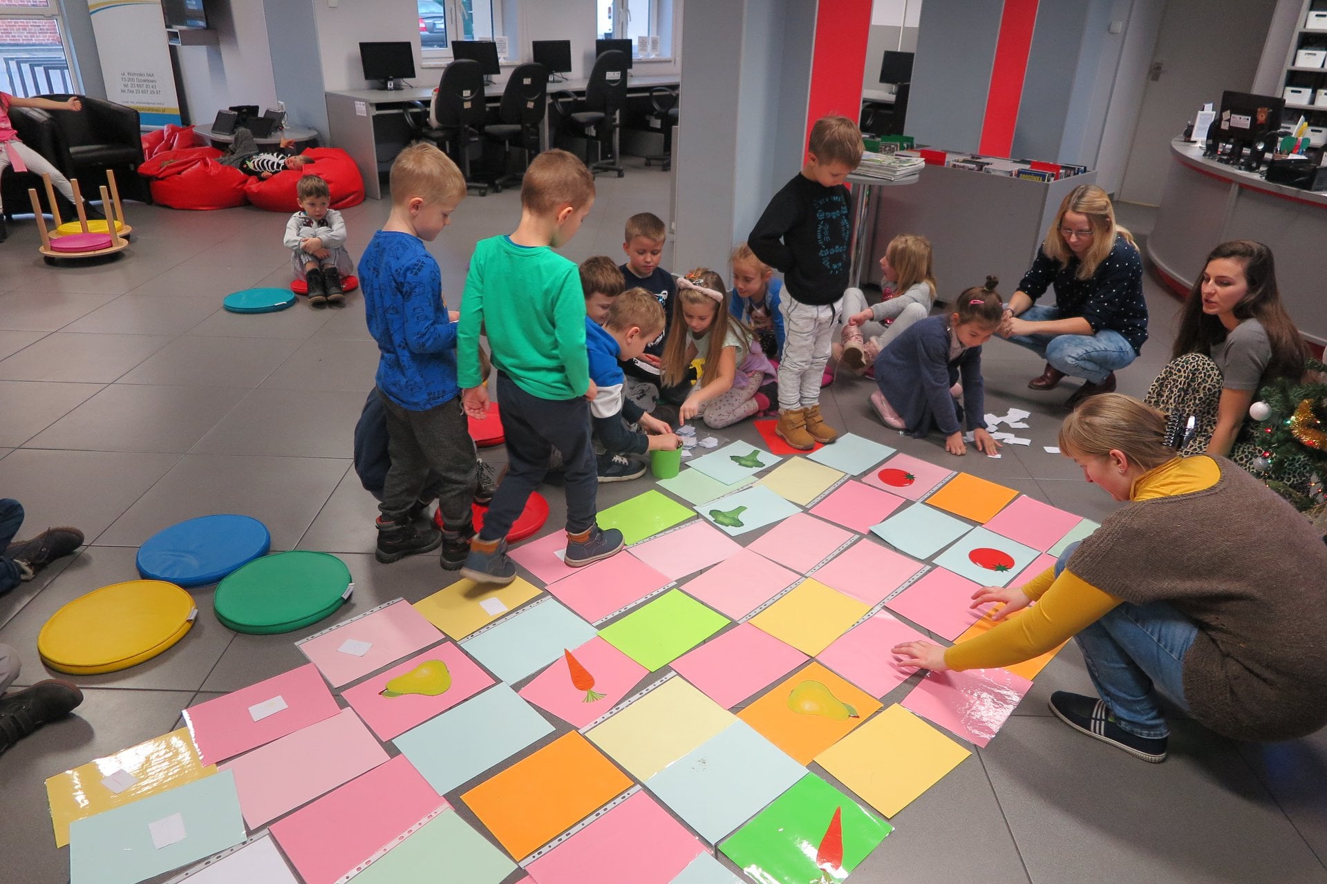 Dzieci z przedszkola "U Misia'' biorą udział w owocowo-warzywnej grze uczącej programowania. Na podłodze rozłożone są kolorowe kartki. Zajęcia prowadzi Małgorzata Trąmpczyńska. 