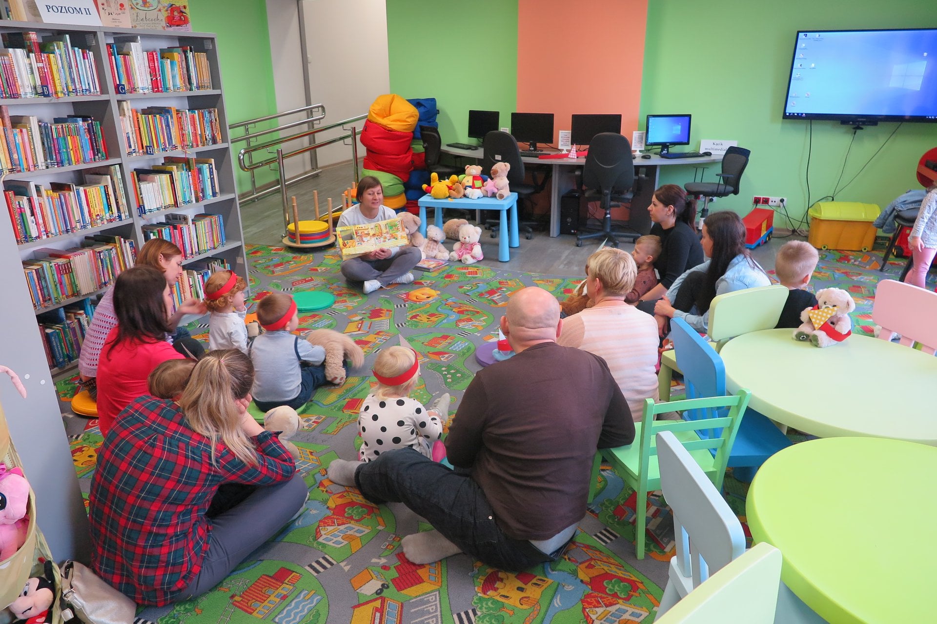 Wnętrze biblioteki (Klub Malucha) dzieci wraz  z opiekunami siedzą w okręgu  i słuchają opowiadania o misiach.Zajęcia prowadzi bibliotekarka Ania Wiśniewska.