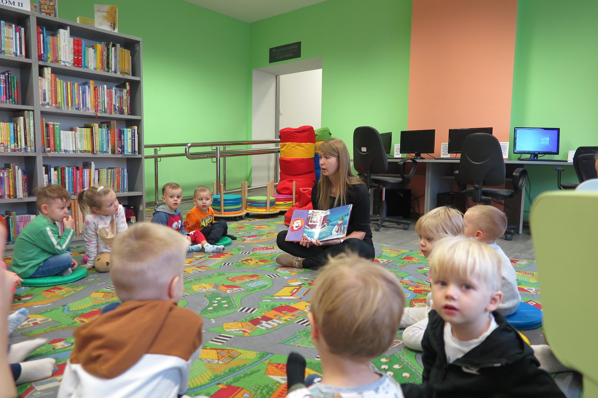 Wnętrze biblioteki. W okręgu siedzą dzieci z przedszkola u Misia grupa (Gumisie). Bibliotekarka Aleksandra Cybulska czyta opowiadanie o liście do świętego mikołaja. 