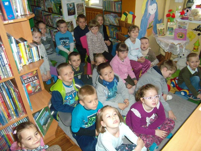Między regałami z książkami siedzi grupa dzieci bierze udział w lekcji bibliotecznej.