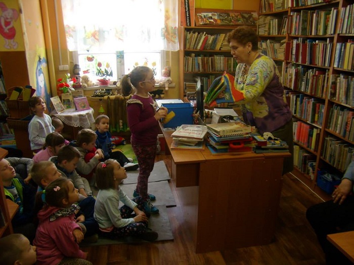 Między regałami z książkami siedzi grupa dzieci bierze udział w lekcji bibliotecznej.