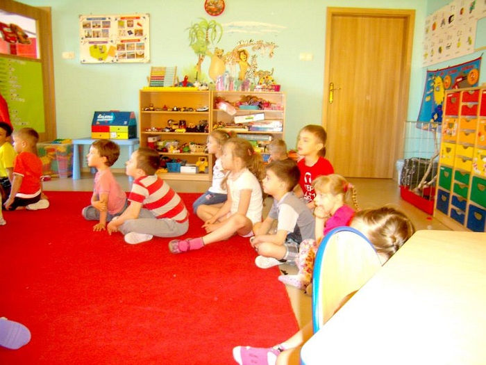 Wnętrze Przedszkola "U Misia'', grupa dzieci siedzi na czerwonym dywanie, w tle zabawki dzieci.