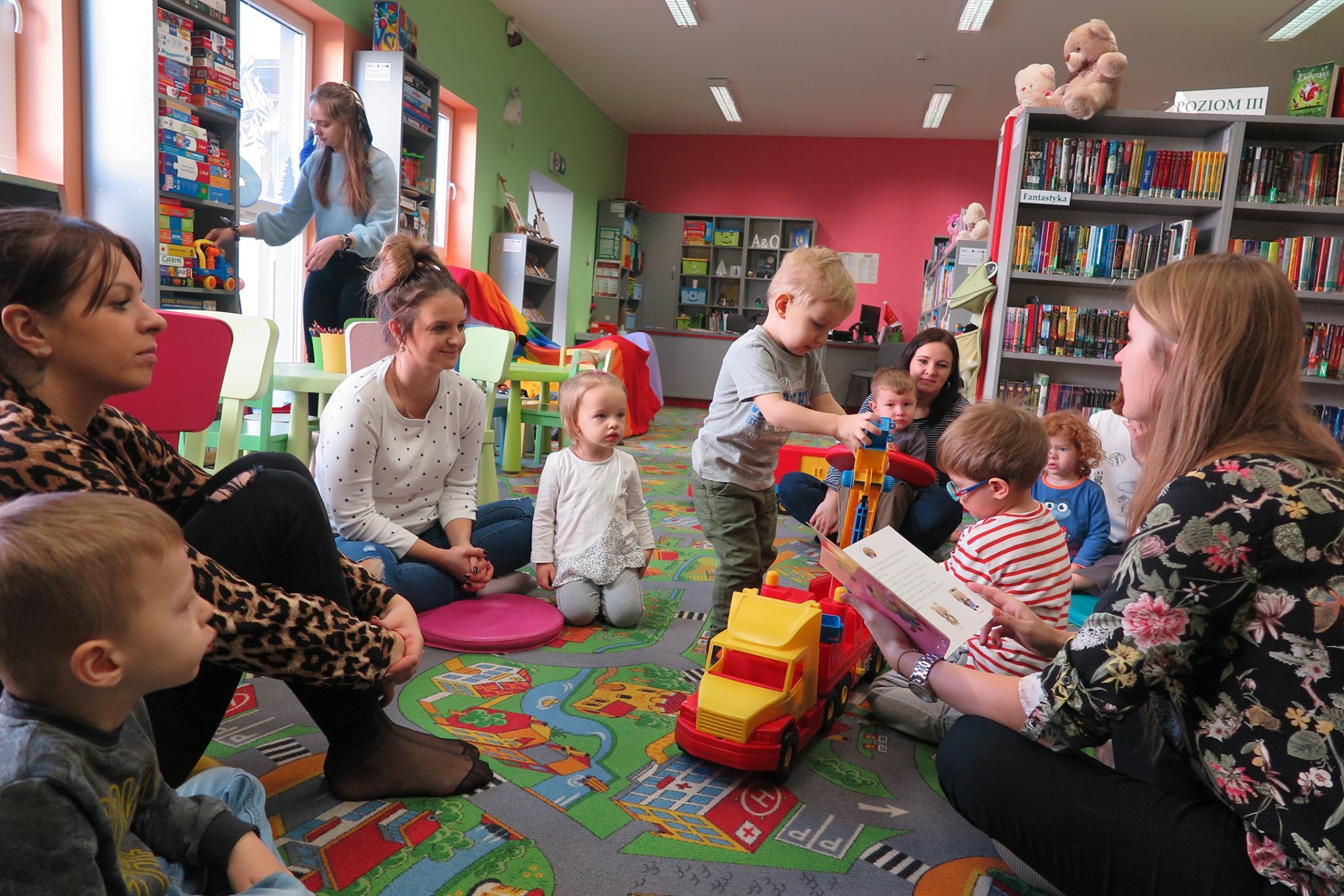 Wnętrze biblioteki, (Klub Malucha) zajęcia prowadzi bibliotekarka Aleksandra Cybulska na temat zbliżającego się święta (Dzień babci i dziadka) , dzieci uważnie słuchaja wraz z rodzicami.
