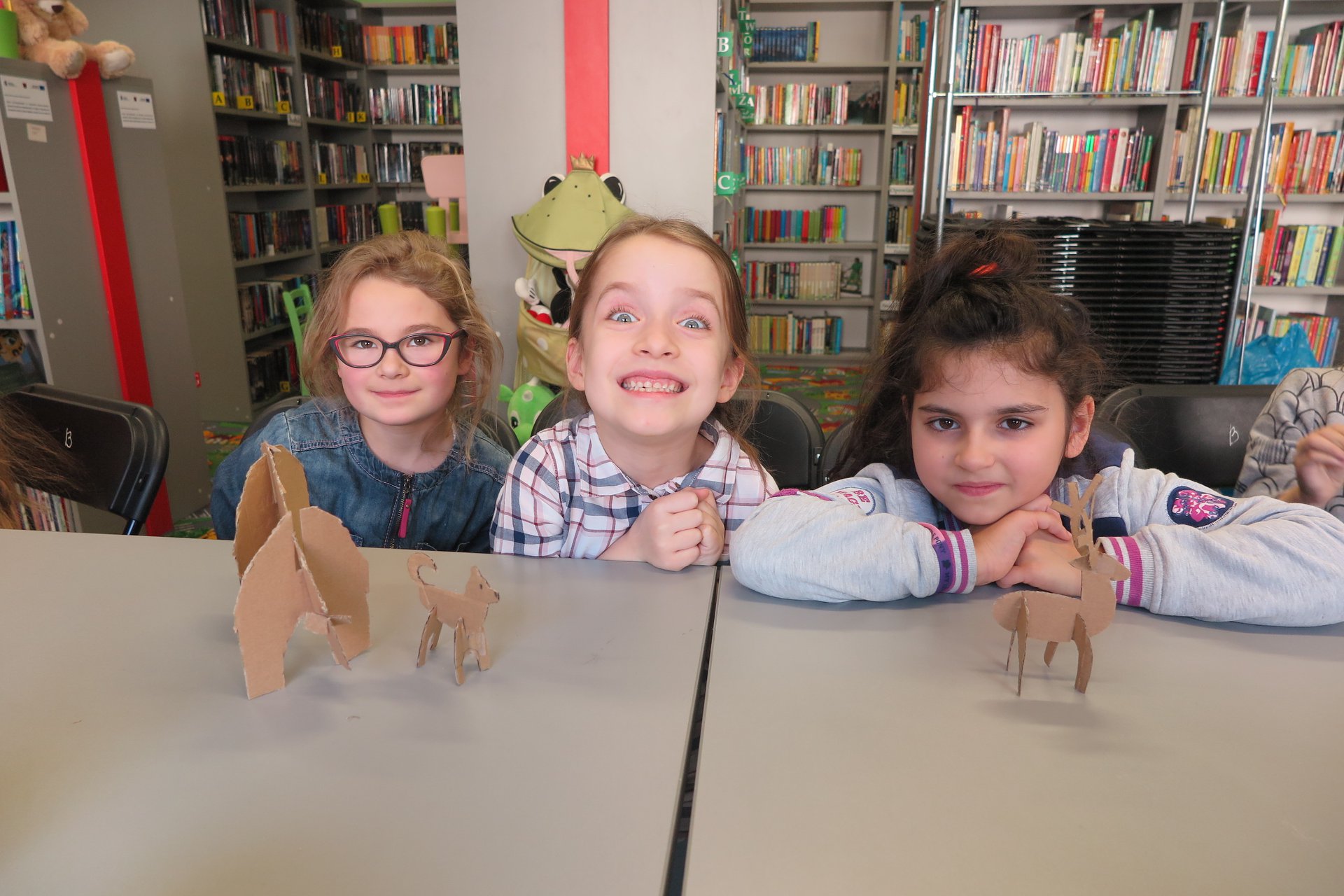 Wnętrze biblioteki,warsztaty plastyczne. Przy stoliku trzy dziewczynki i ich prace wykonane z kartonu. 