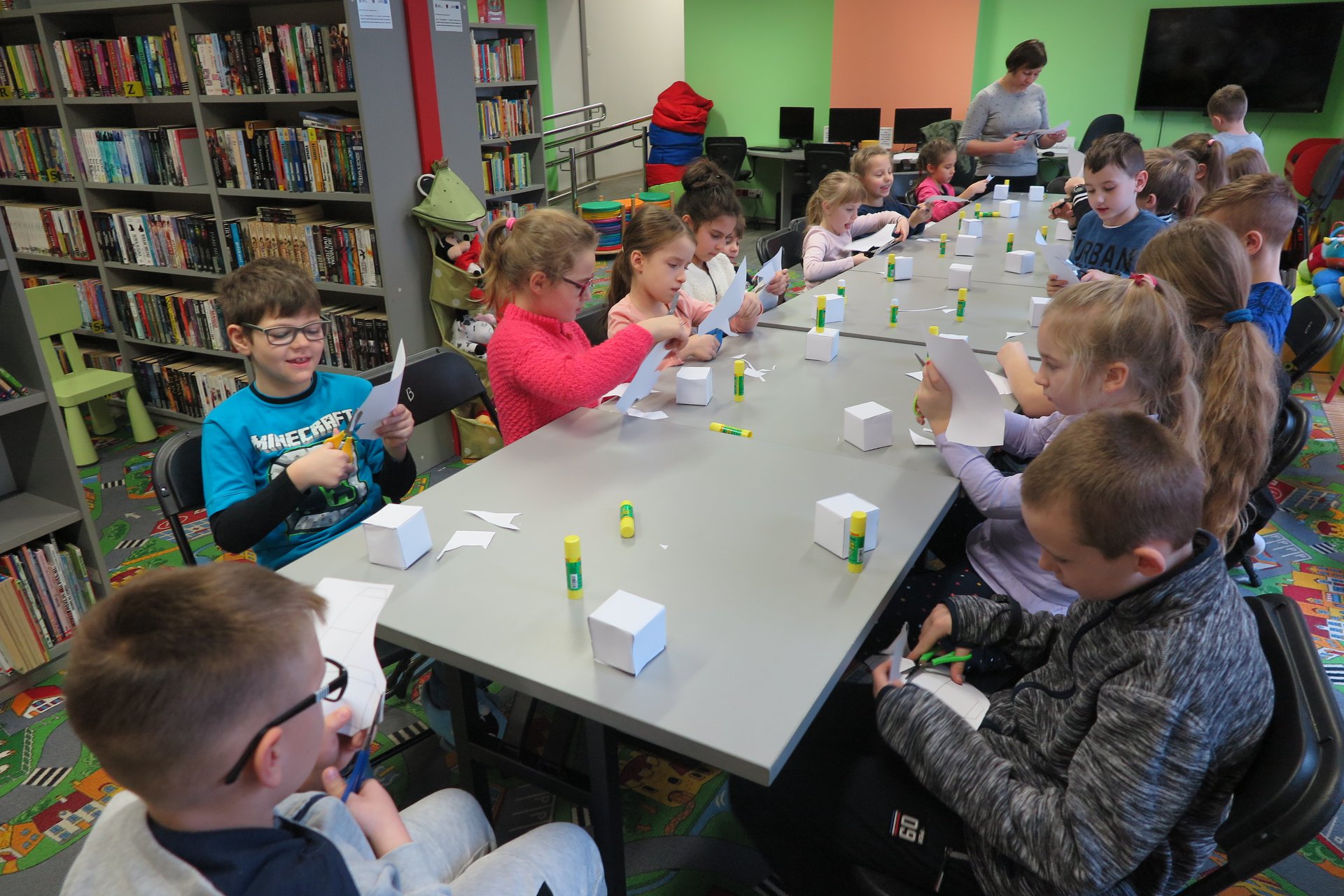 Wnętrze biblioteki przy stolikach siedzą dzieci wykonują pracę z papieru. Zajęcia prowadzi bibliotekarka Ania Wiśniewska.