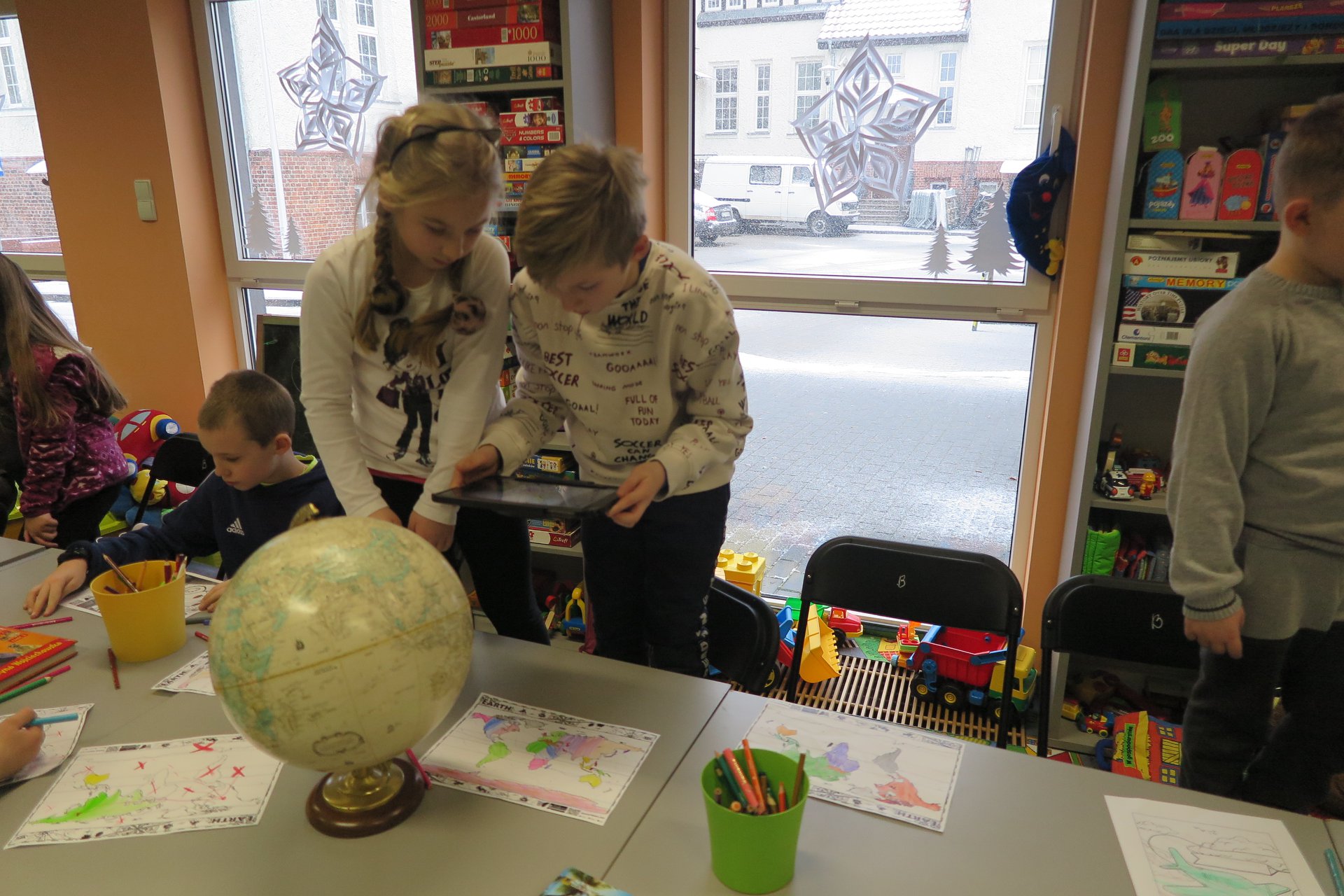 Wnętrze biblioteki (Klub Malucha) warsztaty geograficzne przy stoliku dzieci trzymają tableta i oglądają schematy geograficzne.