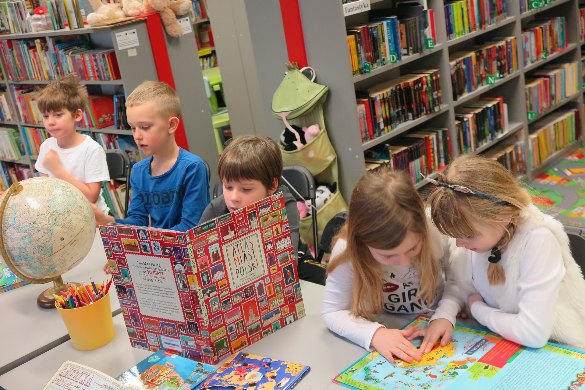 Wnętrze biblioteki (Klub Malucha) warsztaty geograficzne, przy stoliku pięcioro dzieci, chłopiec ogląda atlas miast Polski, dziewczynko obok oglądają mape