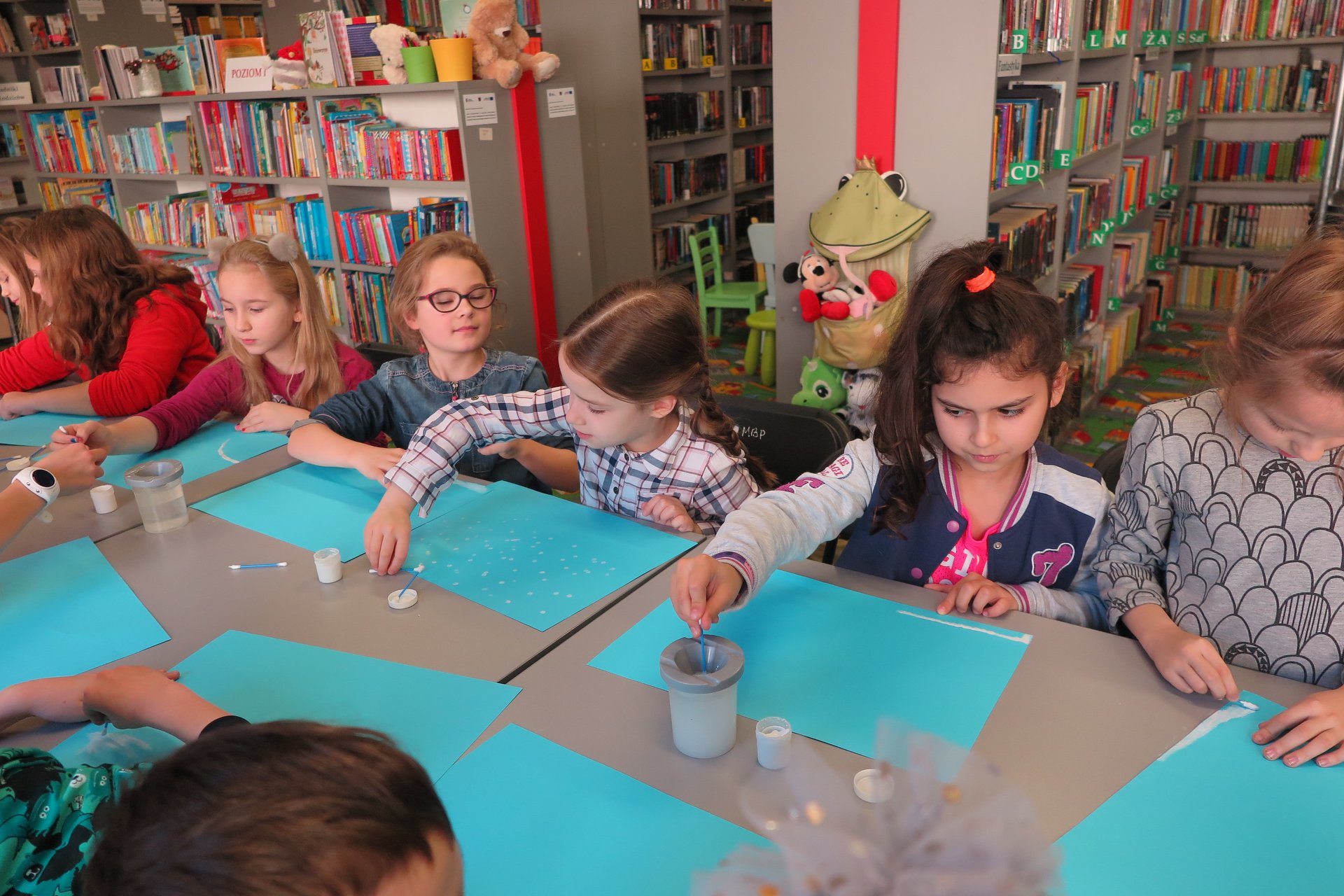 Wnętrze biblioteki,warsztaty plastyczne. Przy stoliku grupa dzieci maluje zimowe krajobrazy. 