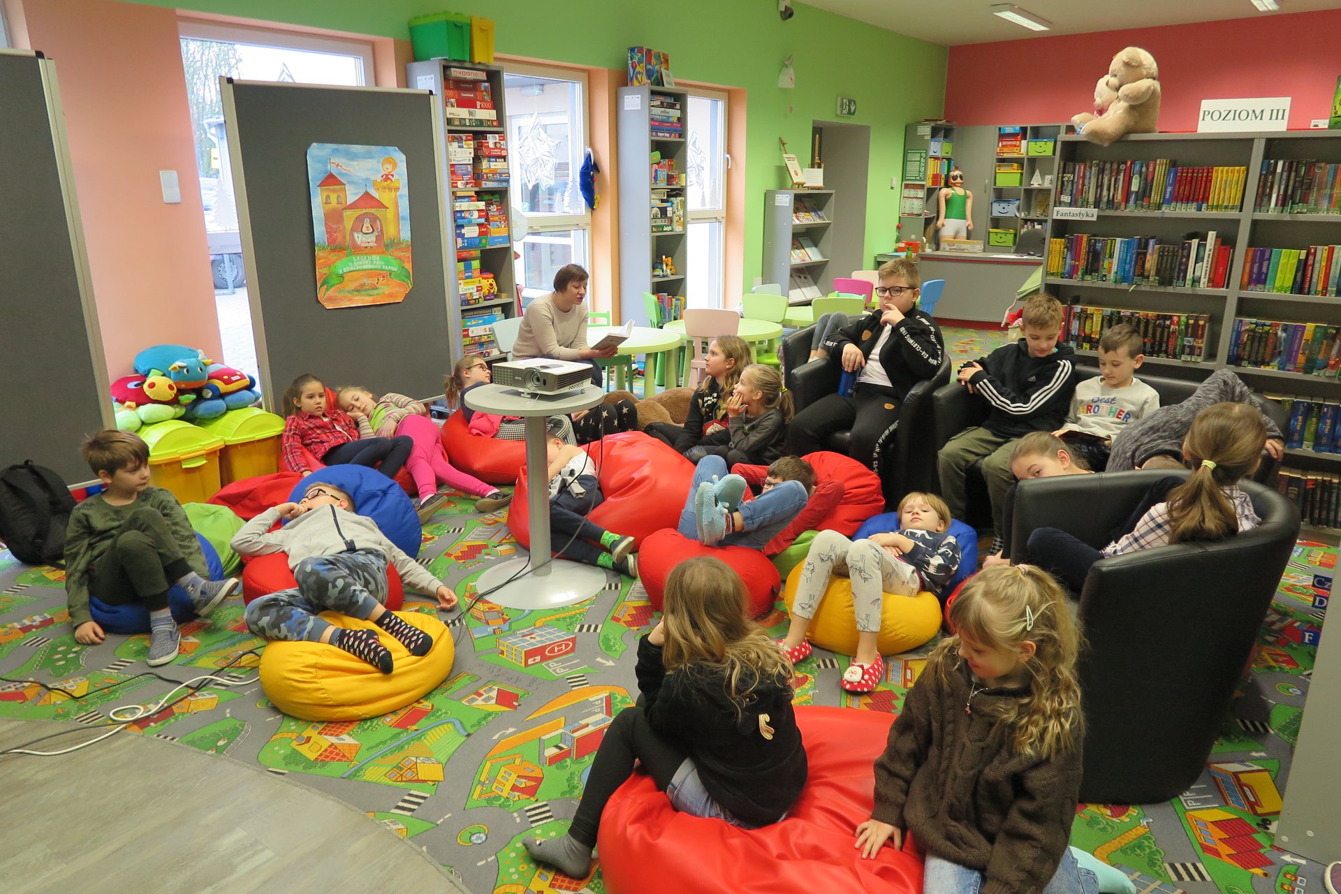 Wnętrze biblioteki, bibliotekarka Ania Wiśniewska czyta dzieciom książkę.
