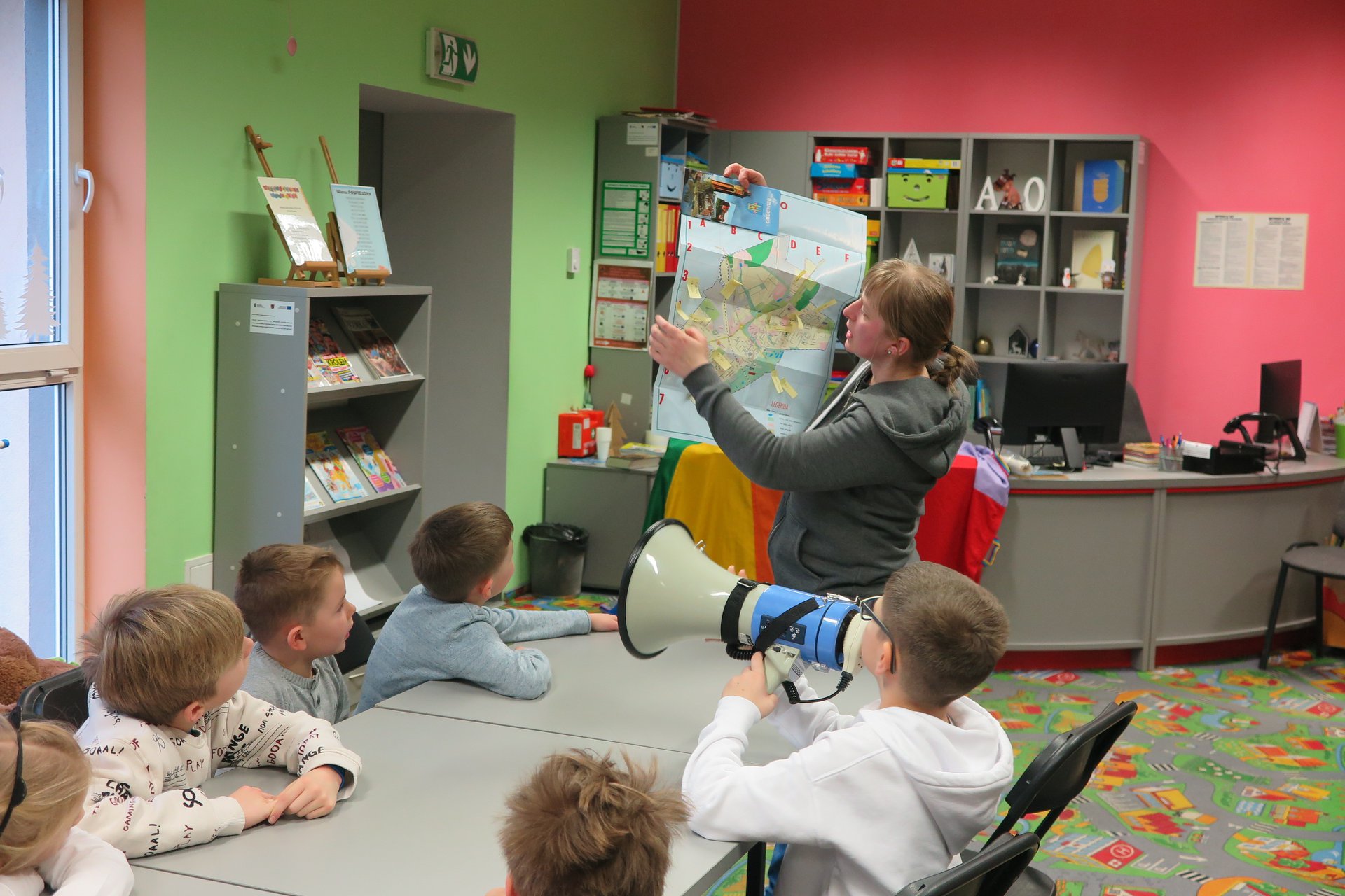 Wnętrze biblioteki (Klub Malucha) warsztaty geograficzne, bibliotekarka Małgorzata Trąmpczyńska prowadzi zajęcia. Przy stolikach siedzą dzieci. Bibliotekarka pokazuje mapę.