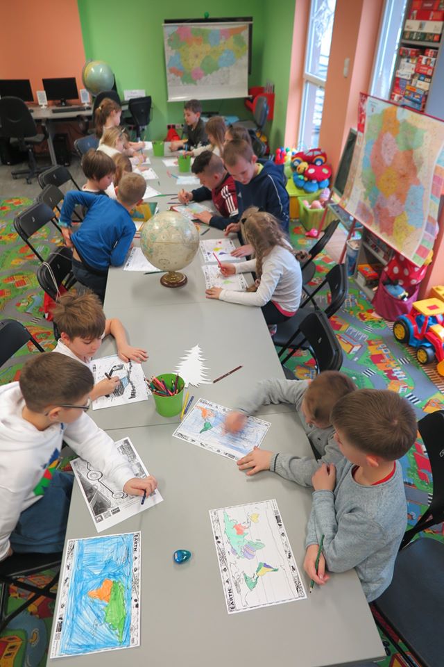 Wnętrze biblioteki (Klub Malucha) trwają warsztaty geograficzne na stole stoi globus dzieci siedzą na krzesełkach i malują mapy.