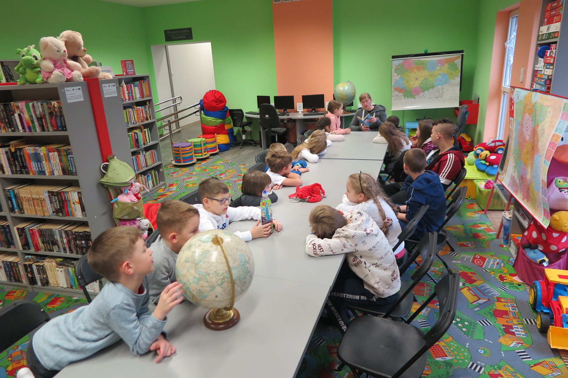 Wnętrze biblioteki (Klub Malucha) warsztaty geograficzne, bibliotekarka Małgorzata Trąmpczyńska prowadzi zajęcia. Przy stolikach siedzą dzieci. Bibliotekarka czyta książkę.