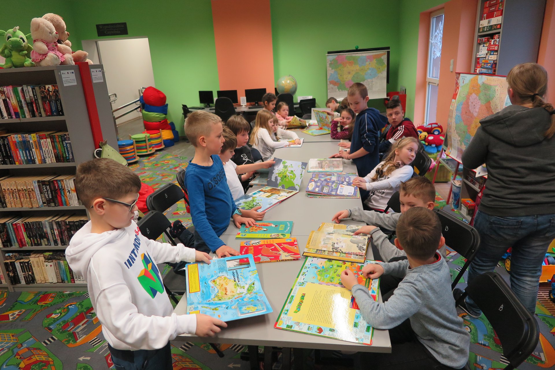Wnętrze biblioteki (Klub Malucha) warsztaty geograficzne przy stolikach siedzą dzieci każde z nich ma książke. Zajęcia prowadzi bibliotekarka Małgorzata Trąmpczyńska. 