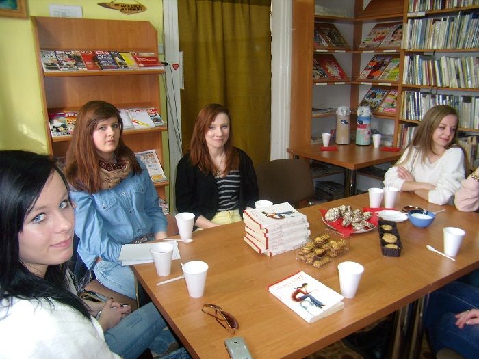 Wnętrze biblioteki, przy stoliku siedzą członkinie DKK wspólnie omawiają  książkę Izabeli Sowy "Agrafka".